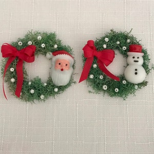 May include: Two miniature Christmas wreaths. Each wreath is made of green moss and decorated with white beads and a red bow. One wreath features a Santa Claus figure, and the other has a snowman figure.