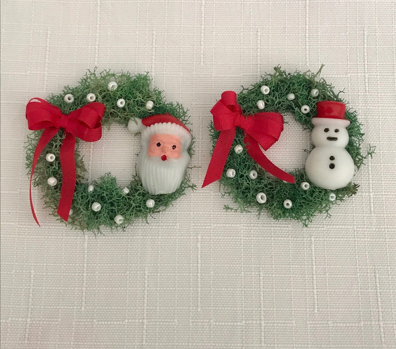 May include: Two miniature Christmas wreaths. Each wreath is made of green moss and decorated with white beads and a red bow. One wreath features a Santa Claus figure, and the other has a snowman figure.