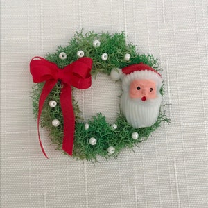 May include: A miniature Christmas wreath with green moss, white pearl accents, and a red ribbon bow. A Santa Claus figure with a red hat and white beard is attached to the wreath. The wreath is a festive holiday decoration.