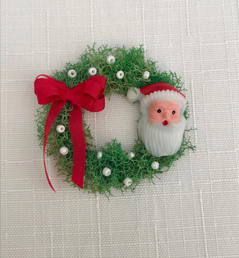 May include: A miniature Christmas wreath with green moss, white pearl accents, and a red ribbon bow. A Santa Claus figure with a red hat and white beard is attached to the wreath. The wreath is a festive holiday decoration.