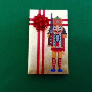 May include: A gold gift box with red ribbon and a nutcracker design. The nutcracker is wearing a red and white uniform with a silver trim and a blue hat with a star.