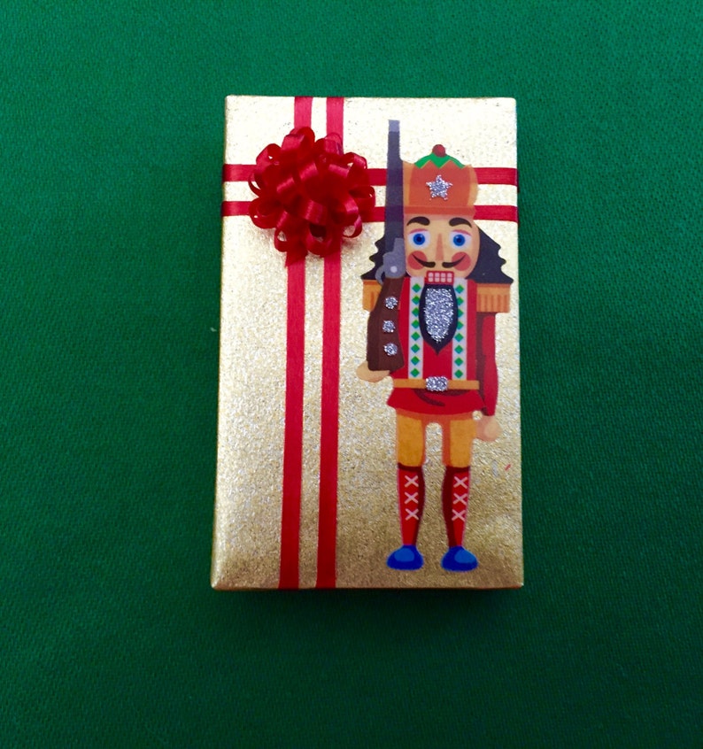 May include: A gold gift box with red ribbon and a nutcracker design. The nutcracker is wearing a red and white uniform with a silver trim and a blue hat with a star.