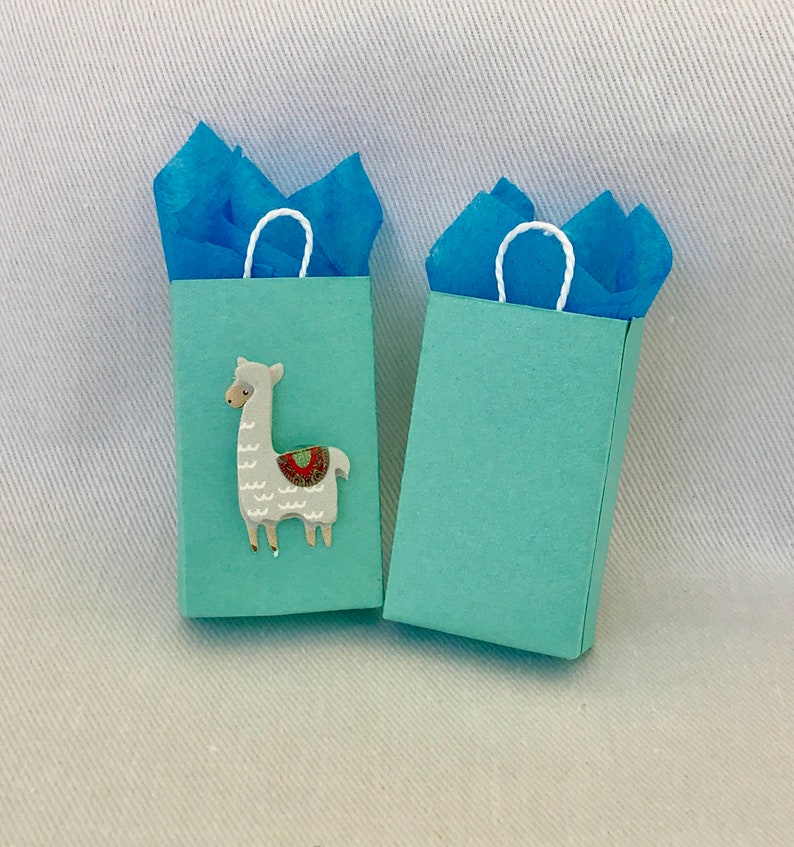 May include: Two light blue gift bags with white handles. One bag features a white llama with a colourful saddle.