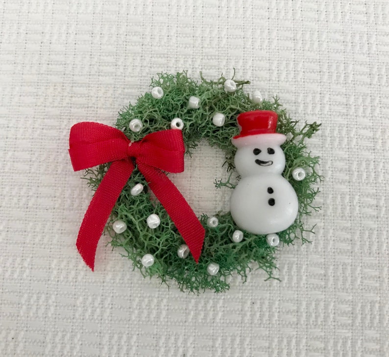 May include: A miniature Christmas wreath made of green moss, adorned with white beads and a bright red bow. A small snowman with a red top hat is featured on the right side of the wreath. Festive holiday decor.