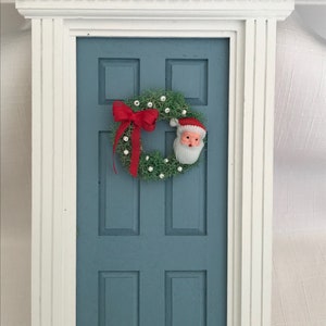 May include: A miniature blue door with a white trim, adorned with a Christmas wreath and a red bow. The wreath features a Santa Claus face and small white ornaments, creating a festive holiday scene.
