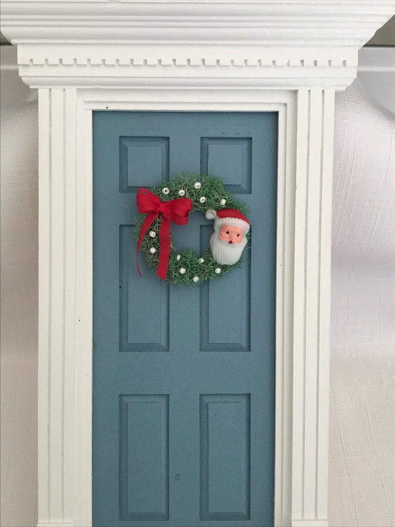 May include: A miniature blue door with a white trim, adorned with a Christmas wreath and a red bow. The wreath features a Santa Claus face and small white ornaments, creating a festive holiday scene.