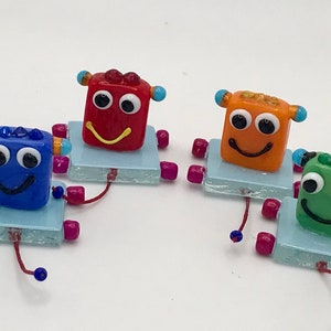 May include: Four small glass robots with smiling faces. Each robot is a different color: blue, red, orange, and green. The robots are on small blue square bases with pink legs.