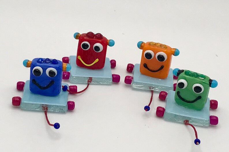 May include: Four small glass robots with smiling faces. Each robot is a different color: blue, red, orange, and green. The robots are on small blue square bases with pink legs.