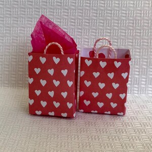 May include: Two small red gift bags with white heart patterns. The bags have white string handles and one bag has pink tissue paper inside.