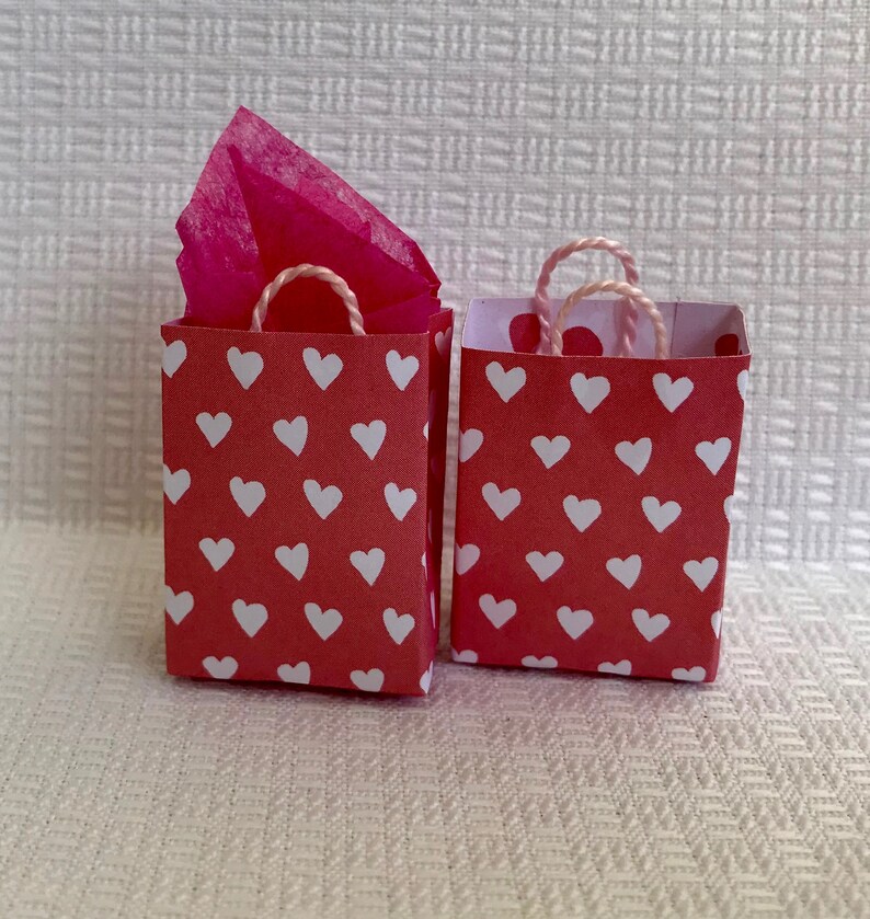 May include: Two small red gift bags with white heart patterns. The bags have white string handles and one bag has pink tissue paper inside.