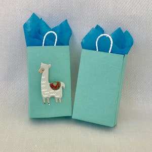 May include: Two light blue gift bags with white handles. One bag features a white llama with a colourful saddle. The bags are filled with blue tissue paper.