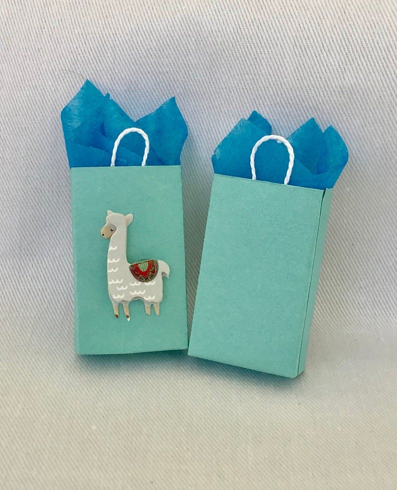 May include: Two light blue gift bags with white handles. One bag features a white llama with a colourful saddle. The bags are filled with blue tissue paper.