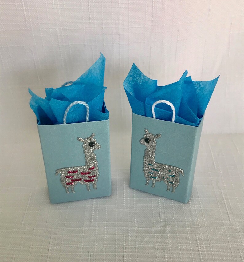 May include: Two light blue gift bags with white handles. Each bag features a silver glitter llama with a different coloured design. The llama on the left bag has a red and white design. The llama on the right bag has a blue and white design.