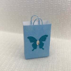 May include: A small light blue gift bag with a turquoise glitter butterfly and a clear rhinestone in the center. The bag has a white string handle.