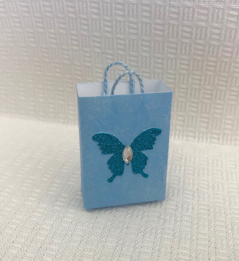 May include: A small light blue gift bag with a turquoise glitter butterfly and a clear rhinestone in the center. The bag has a white string handle.