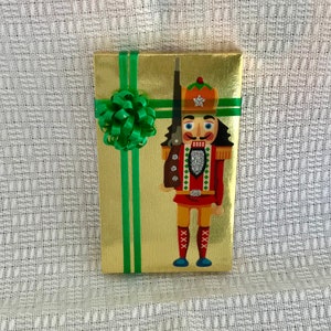 May include: A gold gift box with a green ribbon and bow. The box features a nutcracker illustration wearing a red and green uniform with a white star on his hat.