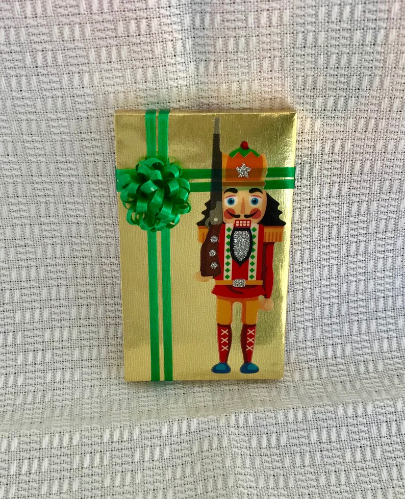 May include: A gold gift box with a green ribbon and bow. The box features a nutcracker illustration wearing a red and green uniform with a white star on his hat.