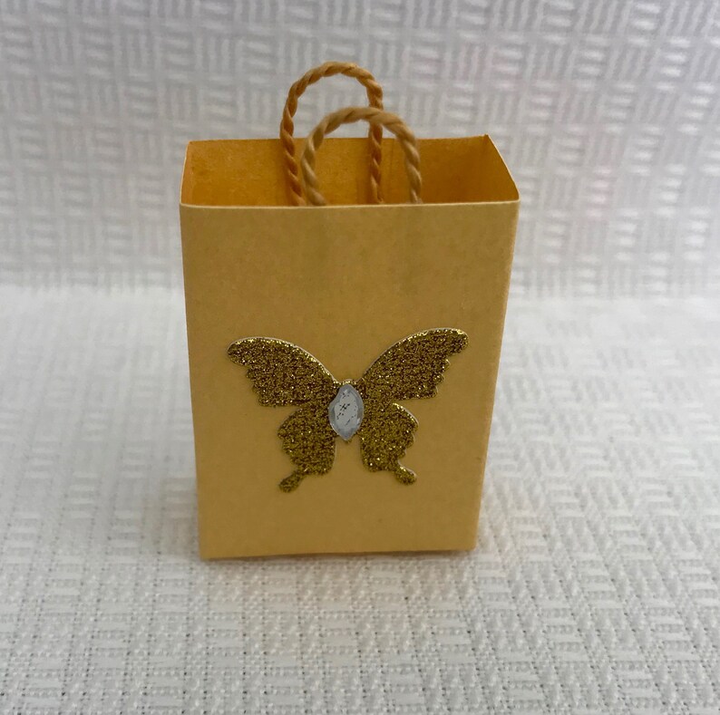 May include: A small, yellow paper gift bag with a gold glitter butterfly design. The bag has a brown rope handle.