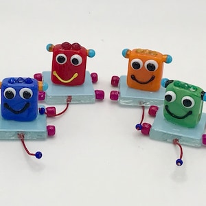 May include: Four small glass robots with smiling faces. Each robot is a different color: red, orange, blue, and green. The robots are on a light blue square base with pink and blue beads for legs.