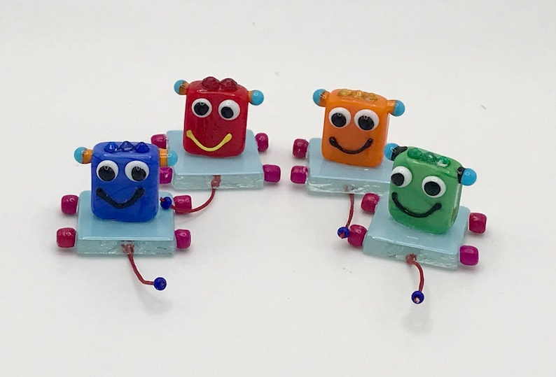May include: Four small glass robots with smiling faces. Each robot is a different color: red, orange, blue, and green. The robots are on a light blue square base with pink and blue beads for legs.
