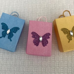 May include: Three small gift bags with butterfly designs. The bags are blue, pink, and yellow. Each butterfly has a rhinestone in the center.
