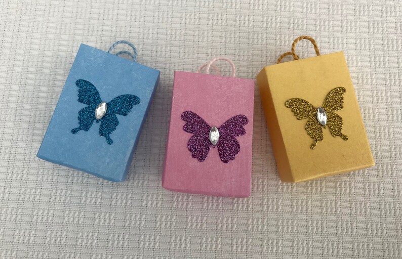 May include: Three small gift bags with butterfly designs. The bags are blue, pink, and yellow. Each butterfly has a rhinestone in the center.