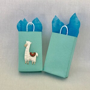May include: Two light blue gift bags with white handles. One bag features a white llama with a colourful saddle.