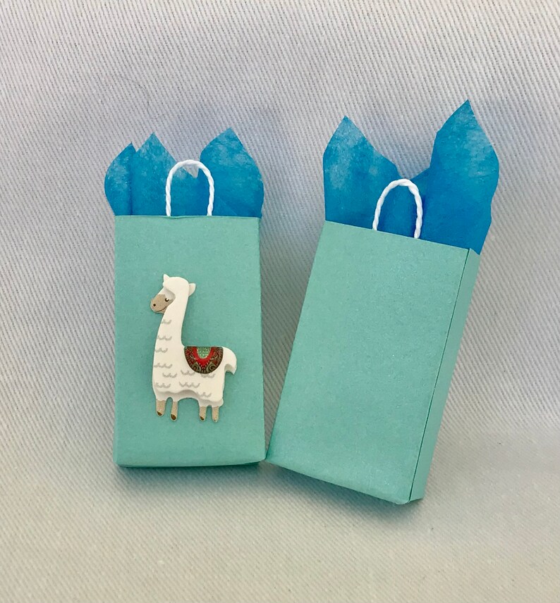 May include: Two light blue gift bags with white handles. One bag features a white llama with a colourful saddle.