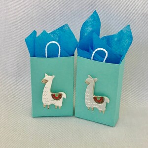 May include: Two small light blue gift bags with white handles. Each bag has a llama cutout on the front. One llama is white with a red and green patterned saddle, and the other llama is grey with a red and green patterned saddle. The bags are filled with blue tissue paper.