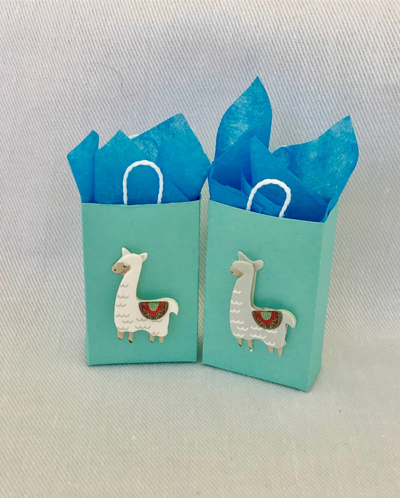 May include: Two small light blue gift bags with white handles. Each bag has a llama cutout on the front. One llama is white with a red and green patterned saddle, and the other llama is grey with a red and green patterned saddle. The bags are filled with blue tissue paper.