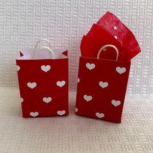 May include: Two red gift bags with white heart patterns and white string handles. The bag on the right has red tissue paper inside.