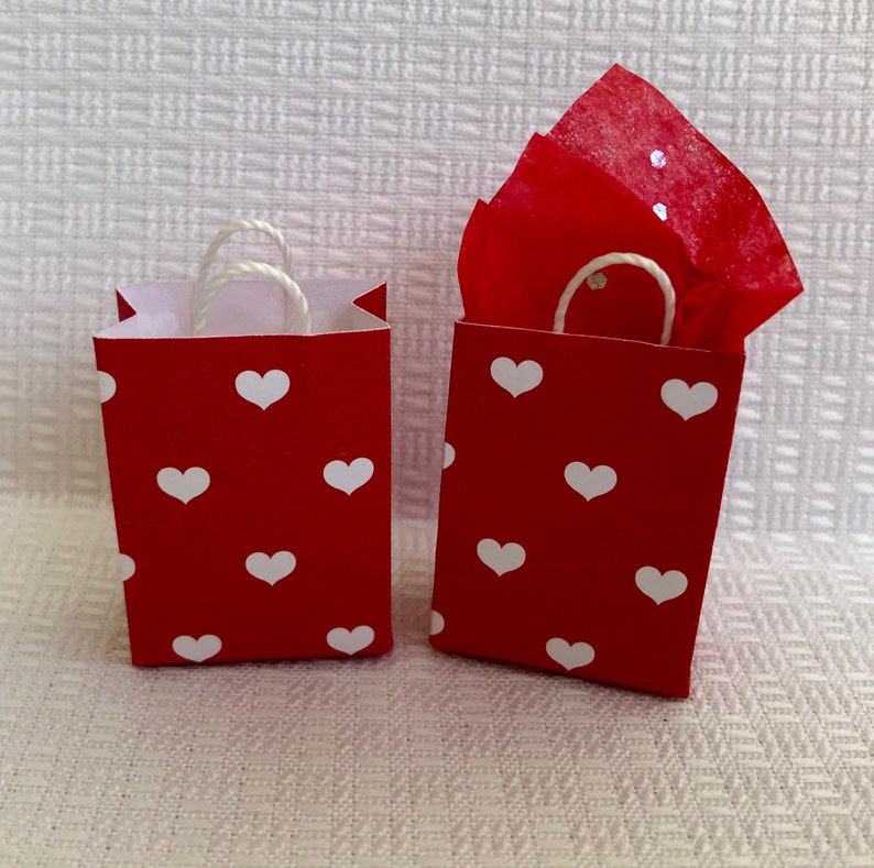 May include: Two red gift bags with white heart patterns and white string handles. The bag on the right has red tissue paper inside.