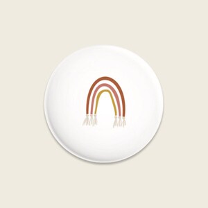 Könnte beinhalten: Ein weißer Keramikteller mit einem Regenbogenmuster. Der Regenbogen besteht aus drei Farben: Rot, Orange und Gelb. Der Regenbogen hat unten einen Fransen.