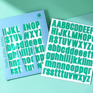 Könnte beinhalten: Eine hellblaue Papphülle mit einem großen ausgeschnittenen Buchstaben M und dem Text Matte Letter Sticker, dazu ein weißer Bogen mit selbstklebenden Alphabet-Aufklebern, die Groß- und Kleinbuchstaben in einem kräftigen Türkisgrün zeigen.