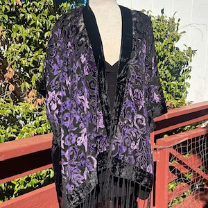 May include: Black velvet kimono with a purple floral pattern. The kimono has a black fringe along the bottom and black velvet trim. The garment is displayed on a mannequin. The background includes greenery and a red wooden deck.