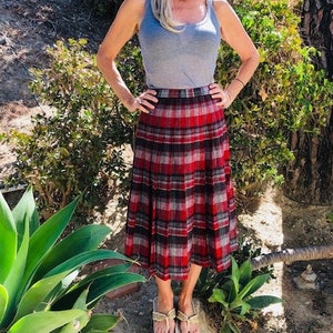 Pode incluir: Uma mulher vestindo um top cinza sem mangas e uma saia xadrez vermelha e preta. Ela está em pé em um jardim.