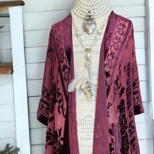 Pode incluir: Um quimono de veludo bordô com estampa floral e franjas. O design aberto drapeia sobre um manequim, mostrando os detalhes e texturas intrincados do tecido. Um colar de pérolas e um pingente adicionam um toque de elegância.