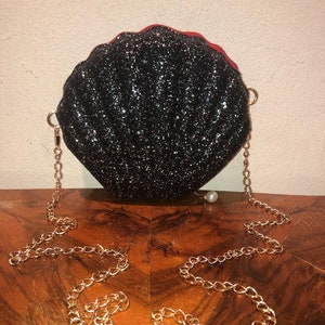 May include: A black glitter seashell-shaped clutch bag with a gold chain strap and a pearl charm.