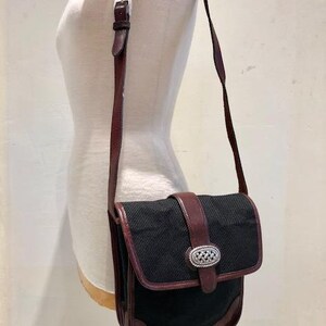 Op de afbeelding: Een zwarte en bruine leren schoudertas met een zilveren gesp. De tas heeft een klep sluiting en een lange verstelbare band.