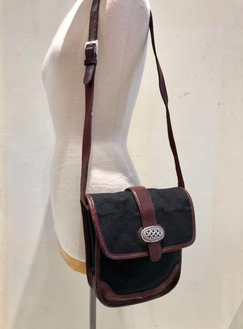 Op de afbeelding: Een zwarte en bruine leren schoudertas met een zilveren gesp. De tas heeft een klep sluiting en een lange verstelbare band.