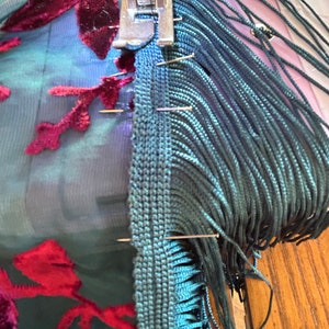 May include: A sewing machine attaching teal fringe to teal fabric with burgundy floral patterns. Pins hold the fringe and fabric in place. The machine's foot is visible, and the work is in progress.