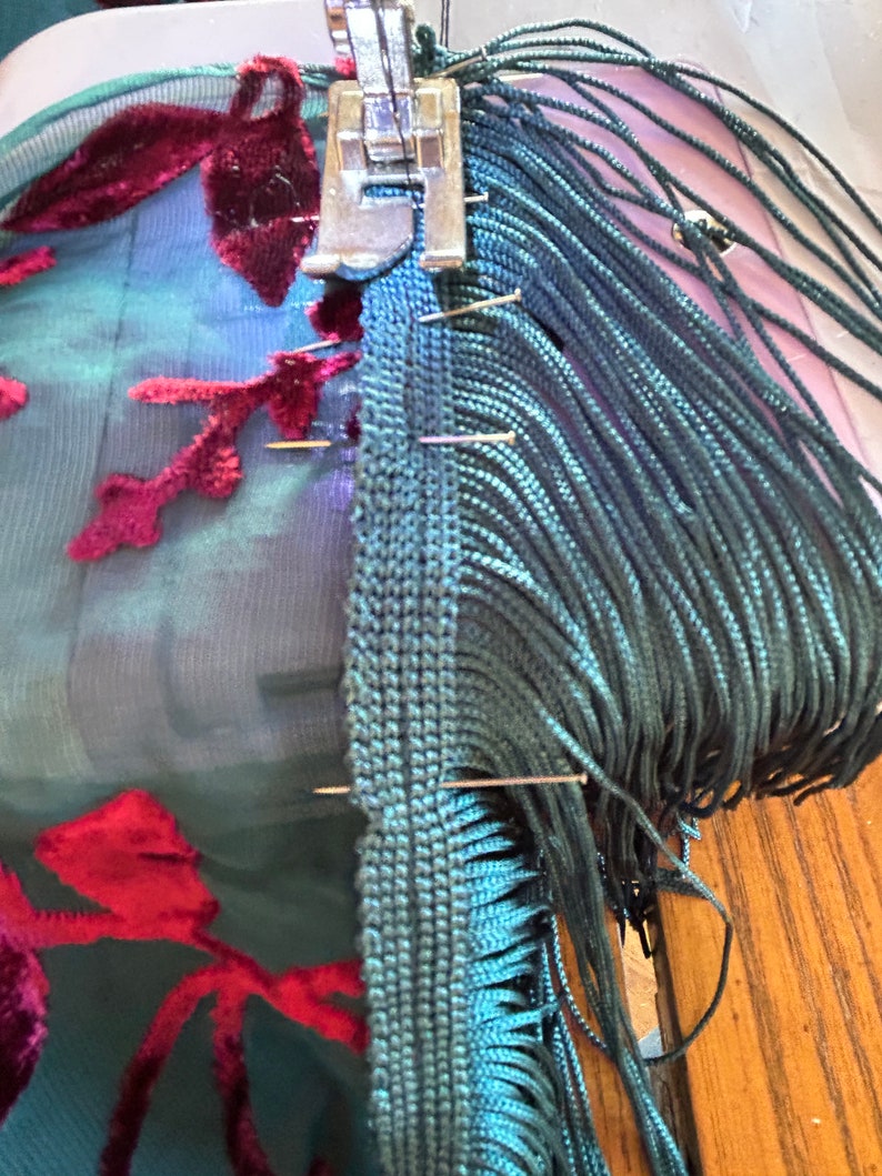 May include: A sewing machine attaching teal fringe to teal fabric with burgundy floral patterns. Pins hold the fringe and fabric in place. The machine's foot is visible, and the work is in progress.