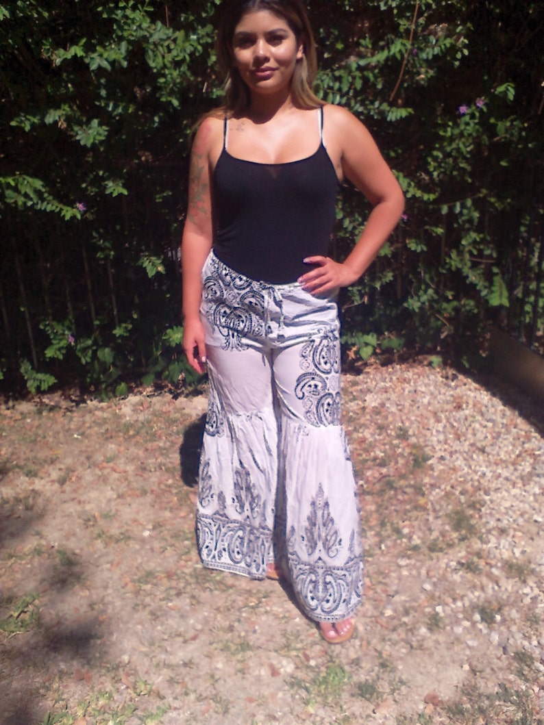 May include: A woman wearing a black tank top and white pants with a black and white paisley pattern. The pants have a wide leg and a ruffled hem.