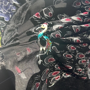 May include: A colorful crane brooch with a white head, turquoise neck, and red, yellow, and black accents. The brooch is set against a black fabric with a velvet peacock feather design, creating a striking contrast.