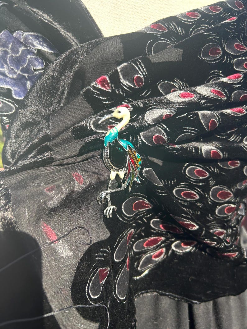 May include: A colorful crane brooch with a white head, turquoise neck, and red, yellow, and black accents. The brooch is set against a black fabric with a velvet peacock feather design, creating a striking contrast.