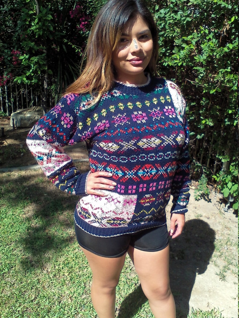 May include: A woman wearing a blue, red, white, and pink patterned sweater with a black and white geometric design. She is also wearing black shorts.