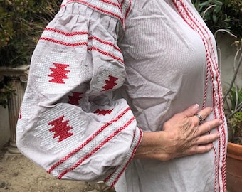 May include: A gray embroidered blouse with red and white stitching. The blouse has a round neckline and long sleeves with a gathered detail at the shoulders. The sleeves are decorated with a geometric pattern.