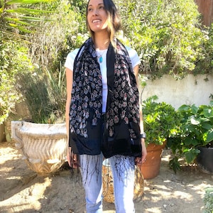 May include: A black scarf with a velvet texture and a peacock feather design. The scarf has a fringe at the bottom and is worn over a white t-shirt and blue jeans. The background includes green plants and a red wall.