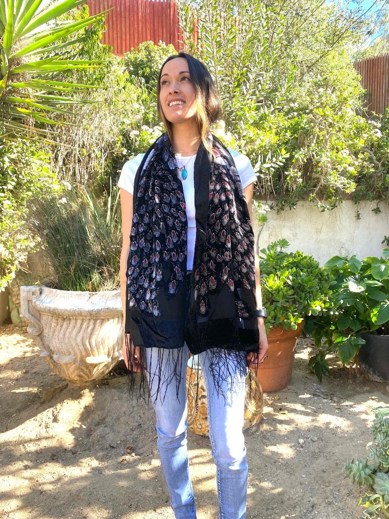 May include: A black scarf with a velvet texture and a peacock feather design. The scarf has a fringe at the bottom and is worn over a white t-shirt and blue jeans. The background includes green plants and a red wall.