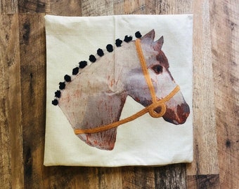 May include: A white pillow with a brown and white horse head design. The horse has a gold bridle and black pom-poms around its mane.